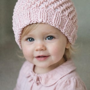 Puede incluir: Un bebé con un gorro de punto rosa claro con un patrón texturizado. El bebé también lleva una camisa con cuello rosa claro. El bebé tiene ojos azules y está sonriendo.