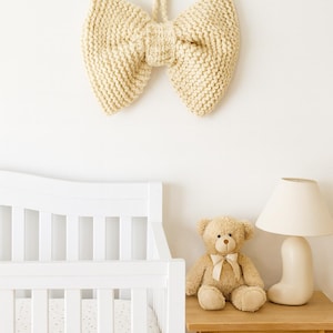 May include: A cream-coloured, knitted bow hangs on a white wall above a white cot. A teddy bear with a cream-coloured bow sits on a wooden side table next to a white lamp. The scene evokes a nursery theme.
