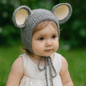 Könnte beinhalten: Ein grauer Babyhut aus Strick mit großen Mäuseohren. Die Ohren haben eine cremefarbene Innenseite. Der Hut wird unter dem Kinn mit grauem Garn gebunden. Das Baby trägt ein weißes Kleid und ist im Freien.