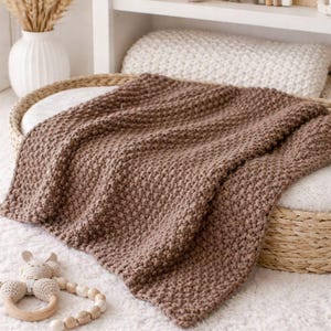 May include: A brown knitted blanket is draped over a white cushion in a wicker basket. A wooden baby rattle and a stuffed bunny are nearby. The blanket has a textured, woven appearance and is a warm, neutral color.