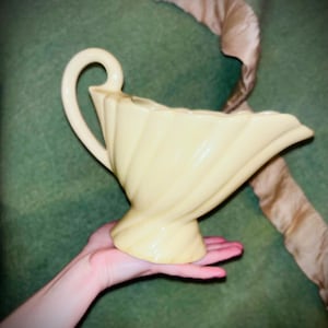May include: A pale yellow ceramic gravy boat featuring a distinct spiral fluted design, a flared pedestal base, and a curved handle. The vessel has a wide, elongated pouring spout and a smooth, glossy finish that reflects light across its surface.