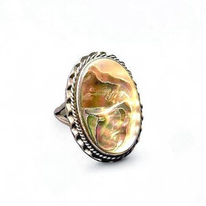 Pode incluir: Anel oval de prata com um grande centro de concha de abalone. A concha iridescente apresenta tons de rosa, verde e dourado. O anel tem uma borda trançada e uma banda simples.