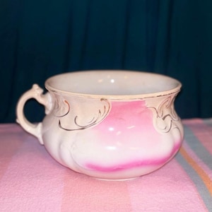 Può includere: Un vaso da notte in ceramica bianca con corpo arrotondato e un manico singolo. L'esterno è decorato con una sfumatura di rosa tenue e delicati motivi a volute color oro lungo il bordo superiore e sui lati del recipiente.