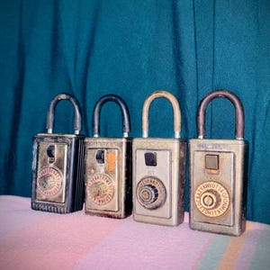 以下が含まれることがあります： 金属製の本体とU字型のシャックルを備えた4つの長方形のSupraキーボックス。各ユニットには中央の押しボタンと、周囲にアルファベットが配置された円形のダイヤルが付いています。仕上げはメタリックな色調でそれぞれ異なります。