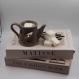 May include: A brown ceramic teapot-shaped oil warmer with leaf cutouts, a lit candle inside, and white wax cubes. The warmer sits atop two books: "MATISSE THE CUT-OUTS" and "THE FASHION BOOK."
