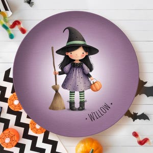 May include: A round, purple plate featuring a cartoon witch holding a broom and a pumpkin. The witch wears a black hat, a purple dress with black polka dots, and green and black striped socks. The name "Willow" is printed on the plate.