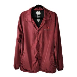 May include: A maroon button-up jacket with a black collar and lining. The jacket has a small red sun graphic and text on the front. The brand name is visible on the collar. The jacket is hanging on a black hanger.