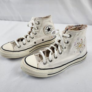 May include: Pair of off-white high-top Converse sneakers. The sneakers feature white laces, a black stripe along the sole, and a black Converse All Star logo patch. One shoe has embroidered sun and star designs.
