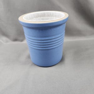 May include: A matte blue ceramic planter featuring a wide, flat beige rim and three horizontal ribbed bands encircling the upper body. The interior of the vessel is white, and the exterior has a smooth, uniform blue finish with a tapered shape.