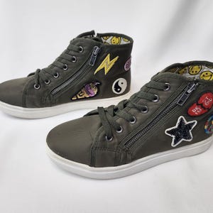 May include: A pair of olive green high-top trainers with white soles. Each shoe has a side zip, laces, and decorative patches, including a lightning bolt, ice cream cone, yin-yang symbol, star, and a broken heart with text.