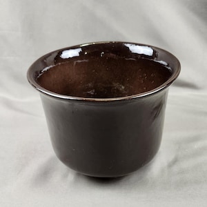 May include: A dark brown ceramic bowl with a smooth, glossy finish and a wide, slightly flared rim. The interior of the bowl is deep and rounded, featuring a consistent dark brown glaze that reflects light across its curved surface.