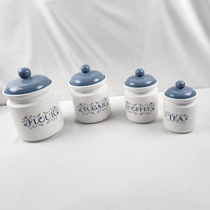 May include: A collection of four white ceramic kitchen storage jars with blue lids. Each jar is labelled with a different word: "FLOUR", "SUGAR", "COFFEE", and "TEA". Floral patterns decorate the jars.