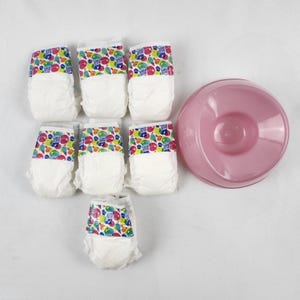 May include: A set of six white baby diapers with colorful patterned waistbands and a pink plastic bowl. The diapers have a design with the words "Baby Alive" and colorful shapes. The bowl is round and has a shallow center.