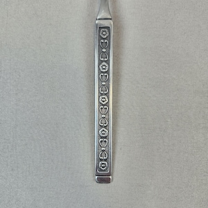 May include: A silver-toned metal utensil handle featuring a vertical decorative pattern of alternating circular flower motifs and small heart-shaped elements. The design is set within a recessed rectangular panel with a textured background finish.