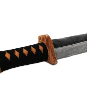 May include: A plush toy sword in a katana style. The blade is gray and black, with a brown guard and handle. The handle is black with diamond-shaped orange accents. The toy is designed for children.