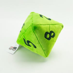 Può includere: Un dado di peluche verde lime a venti facce con numeri neri. Il dado è fatto di materiale morbido e ha i numeri 2, 5 e 8 visibili. Un'etichetta bianca con il testo "GRAFTY" e il logo degli occhiali è attaccata.