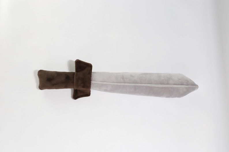 Plush toy sword with a light gray blade and soft brown handle, shaped like a classic medieval weapon. Made from velvety fabric, it's perfect for imaginative play, costumes, or safe mock battles. Handcrafted by the GraTY Foundation – a Polish social enterprise supporting neurodivergent individuals and promoting inclusive employment through creative sewing workshops.