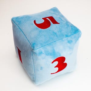 Può includere: Un dado di peluche blu con numeri rossi 1, 3 e 5. Il dado è un giocattolo morbido e coccoloso.