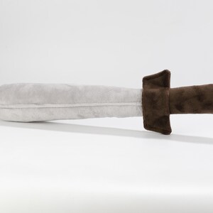 Plush toy sword laying on a flat white surface. The blade is light gray and slightly rounded, made from soft, velvety fabric. The handle and guard are dark brown, offering a secure, cushioned grip. Designed for imaginative play, this handmade sword is part of an inclusive toy line by Fundacja GraTy, a social enterprise supporting neurodivergent makers. Safe, soft, and ideal for young adventurers.