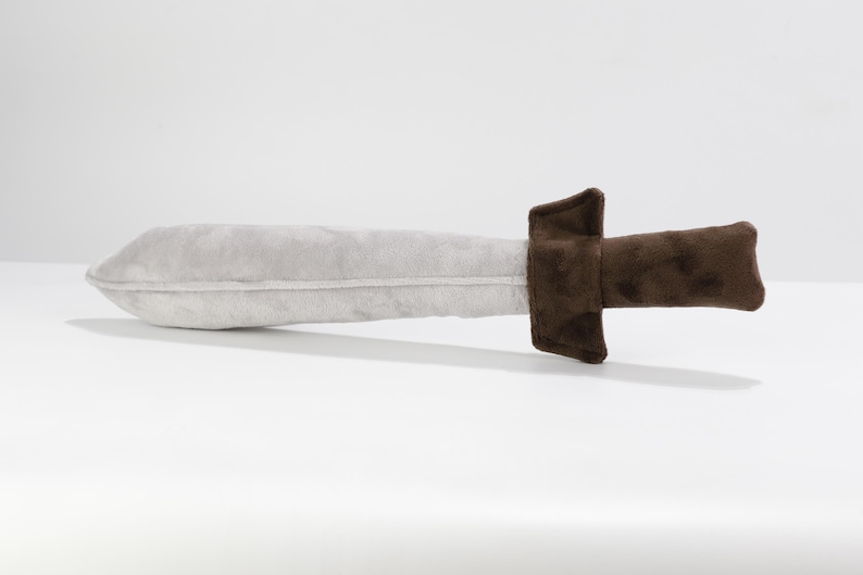 Plush toy sword laying on a flat white surface. The blade is light gray and slightly rounded, made from soft, velvety fabric. The handle and guard are dark brown, offering a secure, cushioned grip. Designed for imaginative play, this handmade sword is part of an inclusive toy line by Fundacja GraTy, a social enterprise supporting neurodivergent makers. Safe, soft, and ideal for young adventurers.