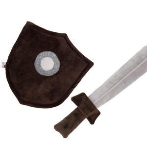 Plush sword and shield toy set on a white background. The sword features a soft gray blade with a brown handle and guard, while the shield is deep brown with a central gray circle and a visible fabric tag. Both items are handmade from soft, velvety materials, ideal for safe, imaginative play. This set is created by Fundacja GraTy, a social enterprise supporting neurodivergent makers and promoting inclusive, therapeutic play experiences.