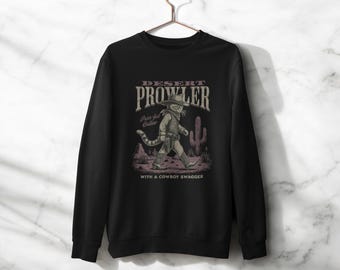 May include: Black long-sleeve sweatshirt with a graphic of a cat dressed as a cowboy walking in a desert scene. The text on the sweatshirt reads "Desert Prowler" and "Pure-feline Outlaw" with the phrase "with a cowboy swagger" below.