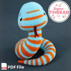 Peut inclure: Un jouet serpent au crochet, rayé de bleu clair et d'orange. La tête est arrondie, avec des yeux noirs et une langue rouge. L'image comprend un logo "Elegant Thread Co" et les mots "PDF File". Un jouet doux et coloré, idéal pour les enfants.