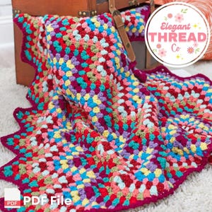 Peut inclure: Une couverture au crochet colorée avec un bord festonné. La couverture est réalisée avec un motif de carrés de grand-mère et présente une variété de couleurs vives, notamment le rouge, le rose, le bleu, le vert, le jaune et le violet. La couverture est pliée et drapée sur un coffre en bois. Le texte "Elegant Thread Co." est visible dans le coin supérieur droit de l'image.
