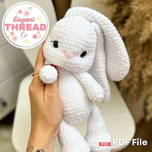 Puede incluir: Un juguete de conejo amigurumi de crochet blanco con orejas largas. El conejo tiene ojos negros y una nariz rosa. El juguete está siendo sostenido por una mano.