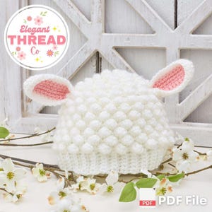 May include: White crocheted baby hat with pink ears, resembling a lamb. The hat is made with a textured stitch, creating a fluffy look. The hat is on a white background with a floral branch and a PDF file icon.
