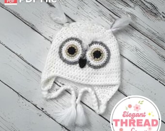 May include: White crocheted owl hat with gray and brown felt accents for the eyes and beak. The hat has a long white tassel and ear flaps. The hat is on a white wood background. The text "Elegant THREAD Co." is on a white circle with pink flowers.