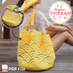 May include: A yellow crochet tote bag with a scalloped design. The bag has a long strap and is made with a delicate open stitch pattern. The bag is shown on a wooden surface with a logo for "Elegant Thread Co." in the background.