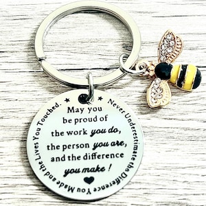 May include: A silver keyring with a round pendant and a bee charm. The pendant has the text "May you be proud of the work you do, the person you are, and the difference you make!" The bee charm is gold, black, and yellow.