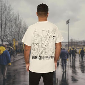 May include: A white t-shirt with a black and white map of Munich, Germany on the back. The map includes the text "MUNICH" and "Allianz Arena 48.2180° N, 11.6347° E".