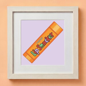 May include: Framed artwork featuring a pixelated orange Lip Smacker lip balm tube. The tube has the words "Lip Smacker" in colorful pixelated text. The artwork is set against a light purple background and framed in a white wooden frame.