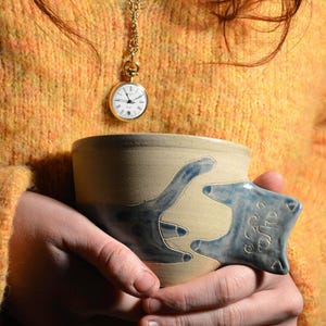 Peut inclure: Une tasse en céramique faite à la main avec une poignée unique en forme de patte de chat, peinte en bleu. La tasse est tenue à deux mains. Une montre de poche dorée sur une chaîne est suspendue au cou. La personne porte un pull jaune.