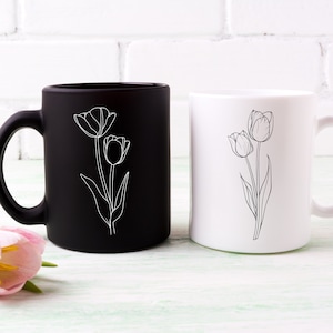 May include: Two ceramic mugs with a simple line drawing of tulips. One mug is black with white tulips, the other is white with black tulips.