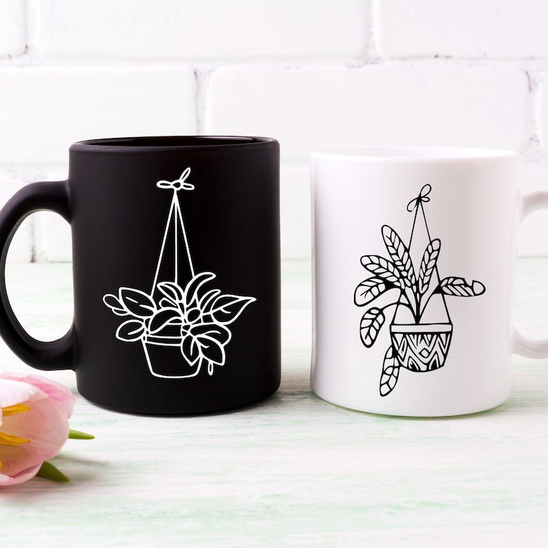 May include: Two ceramic mugs with black and white designs of hanging plants. The mug on the left is black with a white design, and the mug on the right is white with a black design.