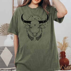 May include: A green t-shirt with a black graphic of a bull's head with a Taurus zodiac symbol and celestial elements.