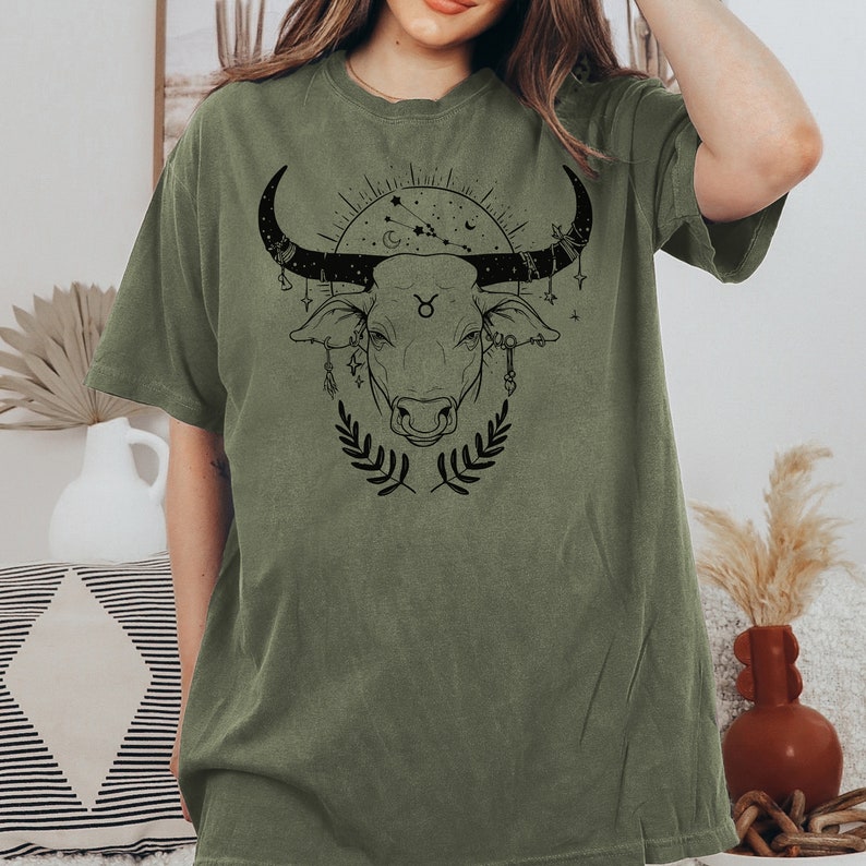 May include: A green t-shirt with a black graphic of a bull's head with a Taurus zodiac symbol and celestial elements.