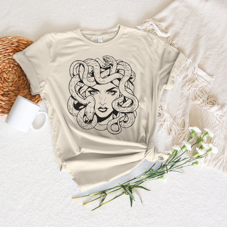 May include: A beige t-shirt with a black and white illustration of Medusa, a mythical Greek figure with snakes for hair.