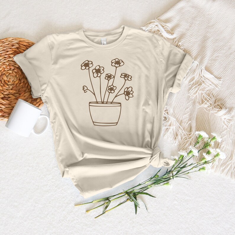 May include: A beige t-shirt with a brown line drawing of a potted plant with flowers. The plant has five flowers with five petals each.