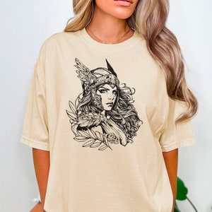May include: A beige t-shirt with a black and white illustration of a woman wearing a helmet with wings and a flowing mane of hair.