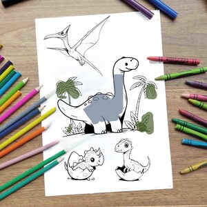 May include: A black and white colouring page featuring a dinosaur, a pterodactyl, and two baby dinosaurs hatching from eggs. The dinosaurs are surrounded by palm trees and green foliage.