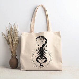May include: A beige tote bag with a black scorpion design. The scorpion is surrounded by stars and a crescent moon. The letter "m" is in the center of the design.