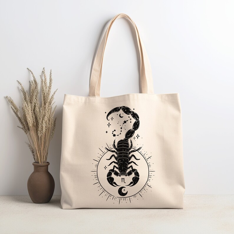 May include: A beige tote bag with a black scorpion design. The scorpion is surrounded by stars and a crescent moon. The letter "m" is in the center of the design.