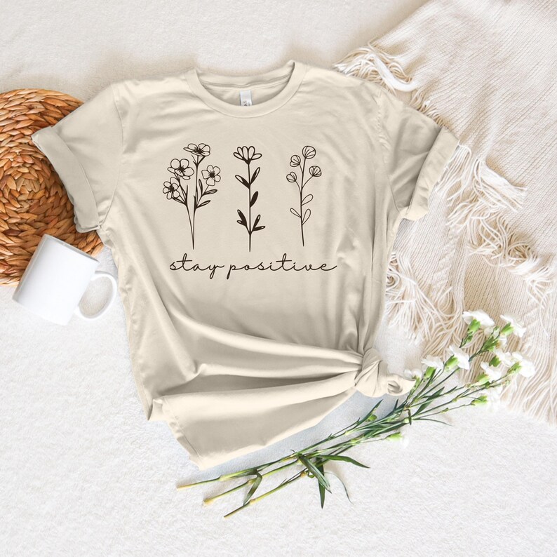May include: A beige t-shirt with a simple design of three stylized flowers and the text "stay positive".