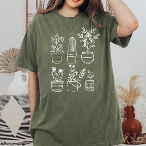 May include: A green t-shirt with a white graphic of potted plants. The plants include a cactus, a succulent, and a variety of leafy plants.