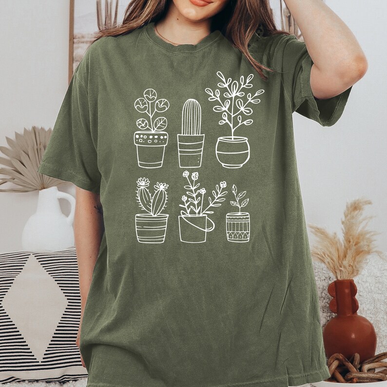 May include: A green t-shirt with a white graphic of potted plants. The plants include a cactus, a succulent, and a variety of leafy plants.
