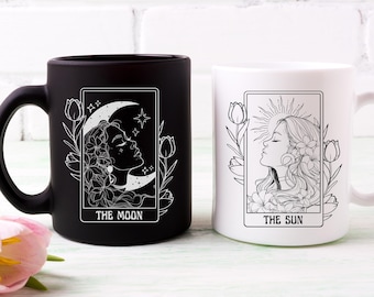May include: Two ceramic mugs with black and white designs. The black mug features a crescent moon and the text "THE MOON". The white mug features a sun and the text "THE SUN".
